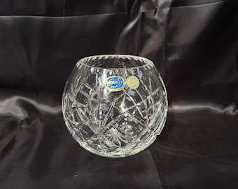 May include: A clear, round crystal bowl with a diamond-cut pattern. The bowl has a wide opening and a label with text. The bowl is sitting on a black surface. This decorative bowl is suitable for home decor.