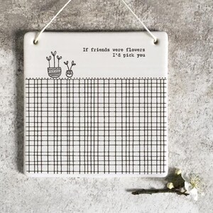Könnte beinhalten: Weiße Keramik-Wandtafel mit schwarzem Gittermuster und dem Text "If friends were flowers I'd pick you". Der obere Teil der Tafel zeigt zwei kleine Topfpflanzen-Illustrationen. Eine Schnur zum Aufhängen ist angebracht.