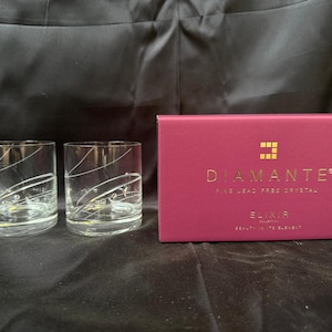 Könnte beinhalten: Zwei klare Kristallgläser mit weißem Wirbeldesign und eine rosa Schachtel mit goldenem Text, der "Diamante Fine Lead Free Crystal" lautet. Auf der Schachtel steht auch "Elixir Beauty With Element".