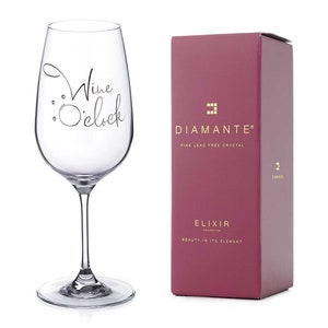 May include: A clear wine glass with the words "Wine O'clock" in a script font. The glass is next to a burgundy box with the "Diamante" logo and "Fine Lead Free Crystal" text. The box also says "Elixir Collection".
