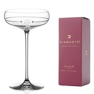 May include: A clear glass champagne flute with a delicate stem and a wide bowl. The bowl is etched with the number "18" and two small dots. The flute is sitting on a white surface. The box in the background is burgundy with gold lettering that reads "DIAMANTE&reg; FINE LEAD FREE CRYSTAL ELIXIR COLLECTION BEAUTY IN ITS ELEMENT".