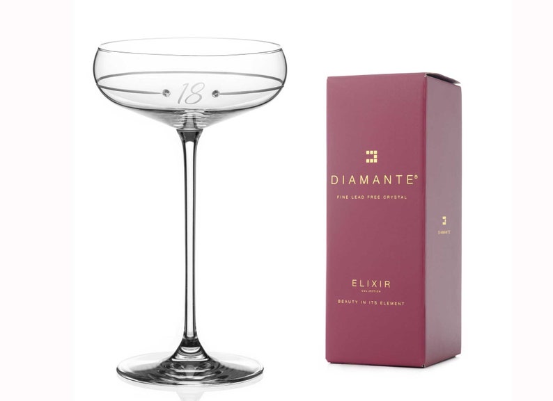 May include: A clear glass champagne flute with a delicate stem and a wide bowl. The bowl is etched with the number "18" and two small dots. The flute is sitting on a white surface. The box in the background is burgundy with gold lettering that reads "DIAMANTE&reg; FINE LEAD FREE CRYSTAL ELIXIR COLLECTION BEAUTY IN ITS ELEMENT".