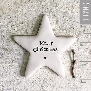 Puede incluir: Un adorno de estrella de cerámica blanca con el texto "Merry Christmas" y un pequeño corazón negro. La estrella mide aproximadamente 3 cm de altura. Una pequeña ramita está al lado de la estrella.