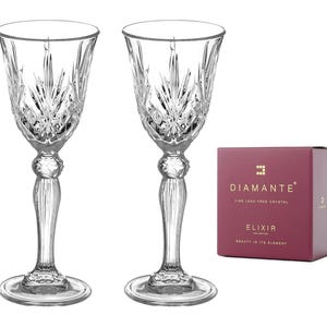 Op de afbeelding: Twee heldere kristallen glazen met een gesneden ontwerp. De glazen staan op een witte achtergrond. De doos op de achtergrond zegt "Diamante Fine Lead Free Crystal Elixir Collections Beauty in its Element"
