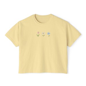 May include: A light yellow t-shirt with a crew neck and short sleeves. The shirt features a small embroidered design of three flowers in pastel colors: pink, white, and blue. The flowers are arranged horizontally across the chest.