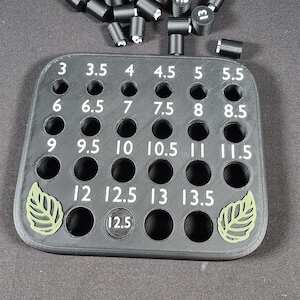 May include: A black, 3D-printed tool with numbered holes for measuring. Cylindrical black pieces with white numbers are scattered around the tool. The numbers on the tool range from 3 to 13.5. Two green leaf designs are on the tool.