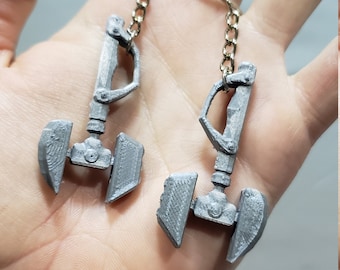 Op de afbeelding: Twee zilverkleurige, 3D-geprinte sleutelhangers in de vorm van een futuristisch wapen met een handvat en een mes. De sleutelhangers zijn bevestigd aan zilveren kettingen.
