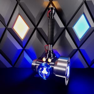 Pode incluir: Um martelo de metal prateado e preto com uma luz azul na frente e duas luzes menores nas laterais. O martelo está em um fundo preto e cinza.