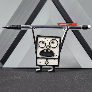 Op de afbeelding: Een wit 3D-geprint beeldje van een cartoonfiguur dat een zwarte en rode pen boven zijn hoofd houdt. Het personage heeft een vierkant lichaam, zwarte ogen en een zwarte mond. Het personage draagt een zwarte stropdas.
