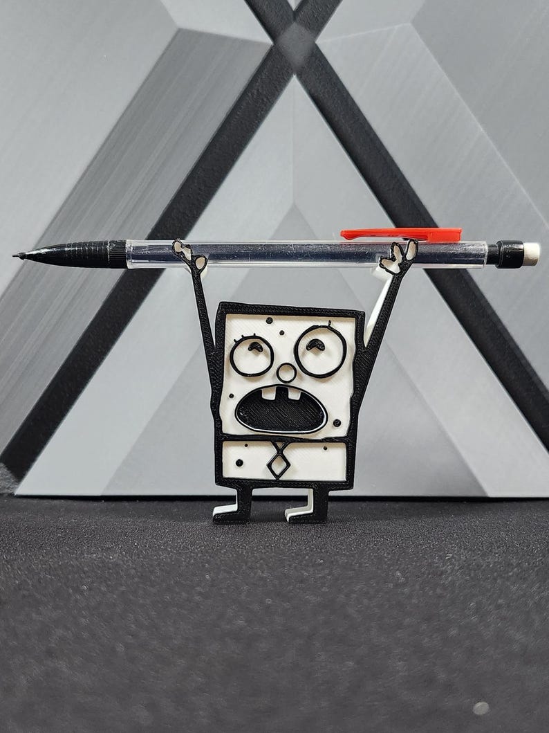 Op de afbeelding: Een wit 3D-geprint beeldje van een cartoonfiguur dat een zwarte en rode pen boven zijn hoofd houdt. Het personage heeft een vierkant lichaam, zwarte ogen en een zwarte mond. Het personage draagt een zwarte stropdas.