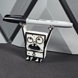 Op de afbeelding: Een wit 3D-geprint beeldje van een cartoonfiguur met zwarte contouren. Het figuur houdt een zwarte en grijze marker met het label "EZZOOL Permanent Marker Fine Tip" boven zijn hoofd.