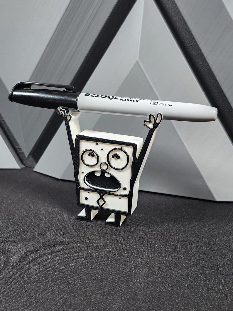 Op de afbeelding: Een wit 3D-geprint beeldje van een cartoonfiguur met zwarte contouren. Het figuur houdt een zwarte en grijze marker met het label "EZZOOL Permanent Marker Fine Tip" boven zijn hoofd.