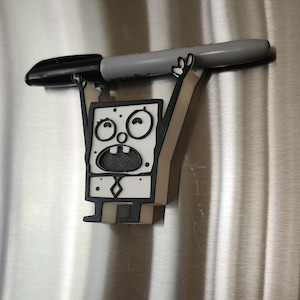 Op de afbeelding: Een 3D-geprint wit en zwart beeldje van een cartoonfiguur met een verraste uitdrukking. Het figuur houdt een grijze marker boven zijn hoofd.