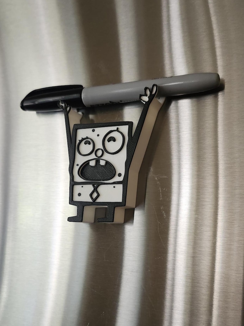 Op de afbeelding: Een 3D-geprint wit en zwart beeldje van een cartoonfiguur met een verraste uitdrukking. Het figuur houdt een grijze marker boven zijn hoofd.