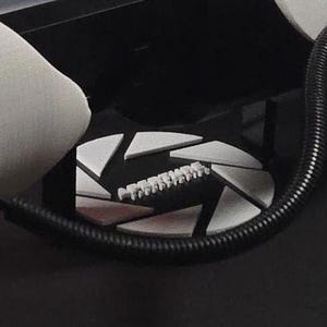 May include: A black and white camera lens cap with a shutter design. The lens cap has a row of small white teeth along the edge.