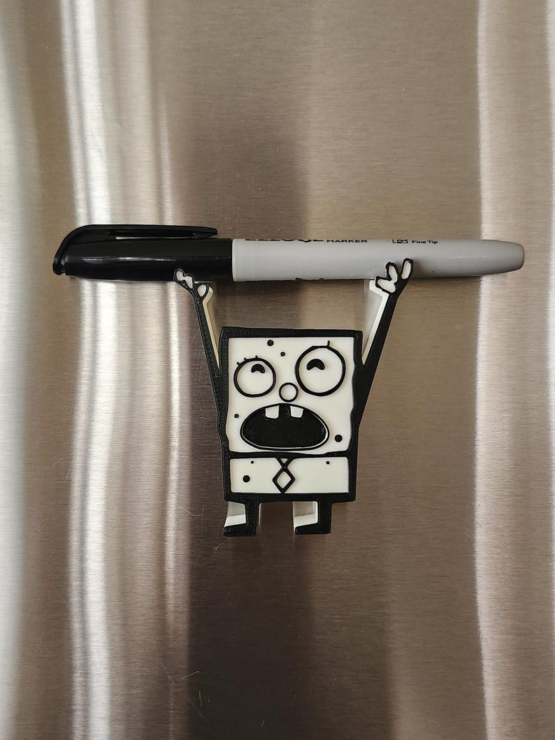 Op de afbeelding: Een wit 3D-geprint beeldje van een cartoonfiguur met zwarte contouren. Het personage heeft een brede mond, grote ogen en houdt zijn armen in de lucht. Het beeldje is bevestigd aan een zilveren koelkast met een zwarte marker.