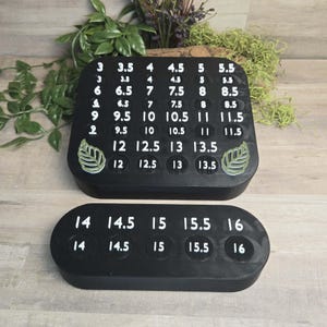 May include: Two black shoe size guides with white numbers. The top guide is rectangular with sizes 3 to 13.5. The bottom guide is oval with sizes 14 to 16. Both guides have leaf accents.