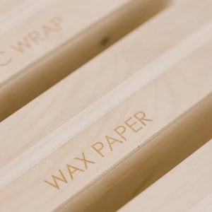 May include: Close-up of a wooden organizer with two slots labeled "Wax Paper" and "Wrap".