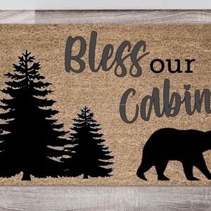 May include: A brown coir doormat with the text "Bless our Cabin" in gray lettering. The doormat features a black silhouette of a bear walking to the right and two pine trees on the left.