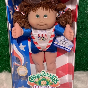 May include: A Cabbage Patch Kids OlympiKids Special Edition doll, Stacy Belinda, in a clear plastic box. The doll wears a red, white, and blue gymnastics uniform with the USA Olympic team logo. The box features patriotic colors and the OlympiKids logo.