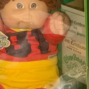 Puede incluir: Una muñeca Cabbage Patch Kids en una caja. La muñeca tiene cabello castaño rizado, una cara redonda y viste una camiseta roja y negra con el número 31. La muñeca también lleva pantalones cortos amarillos. La caja incluye un certificado de nacimiento.