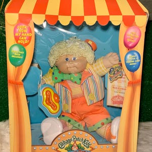 May include: A Cabbage Patch Kids Circus Kids doll dressed as a clown. The doll has blonde curly hair, a red nose, and is wearing a colourful striped shirt, orange trousers, and a yellow and green polka dot waistcoat. The doll is holding a red and yellow toy horn. The doll is packaged in a box with a circus tent design. The box also includes a bonus poster and trading card.