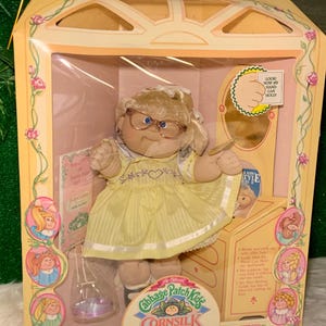 May include: A Cabbage Patch Kids Cornsilk Kids doll in a yellow dress with a heart on the front. The doll is wearing glasses and has blonde hair. The doll is in a box with a pink and white floral design. The box also includes a small mirror and a comb.