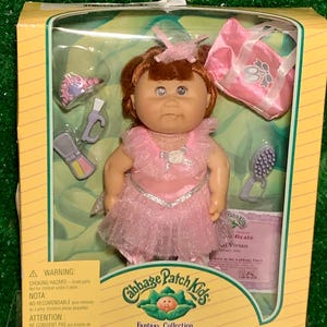May include: Cabbage Patch Kids Fantasy Collection doll, Vivian, in original packaging. The doll has red hair and wears a pink dress with a silver trim. Accessories include a pink purse, shoes, and a brush. The box has a warning label.
