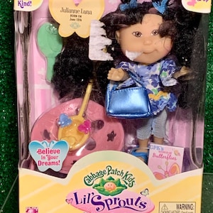 Puede incluir: Muñeca Cabbage Patch Kids Lil' Sprouts con cabello negro y un vestido floral azul y blanco. La muñeca sostiene un bolso azul y un libro que dice "CPK's World of Butterflies". La muñeca está sentada en un juego de niños rosa y amarillo con una pala amarilla y un cubo rosa.