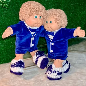 May include: Two Cabbage Patch Kids dolls wearing matching blue velvet outfits with white trim and bow ties. They are both wearing white and purple crocheted shoes.