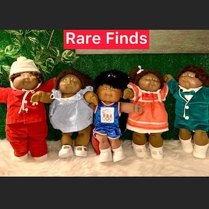 Może przedstawiać: Pięć lalek Cabbage Patch Kids, wszystkie z brązową skórą i czarnymi włosami. Lalki są ubrane w różne stroje, w tym czerwony kombinezon, niebieską sukienkę, czerwoną i białą sukienkę, niebiesko-biały strój sportowy i zielony garnitur. Lalki są ustawione w rzędzie na białym, puszystym podłożu. Tekst "Rare Finds" jest wyświetlany nad lalkami.