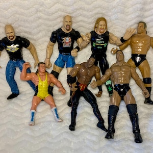 May include: A collection of seven wrestling action figures, including a wrestler with a yellow and black wrestling outfit, a wrestler with a black and green wrestling outfit, a wrestler with a black and red wrestling outfit, a wrestler with a black and blue wrestling outfit, a wrestler with a black and white wrestling outfit, a wrestler with a black and red wrestling outfit, and a wrestler with a black and white wrestling outfit.