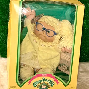 Może przedstawiać: Lalka Cabbage Patch Kid ubrana w żółty sweter i sukienkę z parą żółtych bucików. Lalka ma blond włosy i nosi okulary. Lalka znajduje się w pudełku z tekstem "1985 Cabbage Patch Kids" i "Jestem wyjątkowa. Jestem adoptowalna!" Pudełko jest żółte z zielonymi obwódkami.