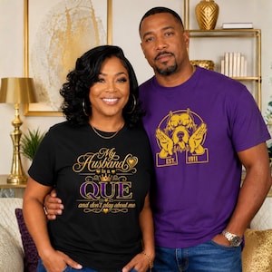 May include: A black t-shirt with gold and purple text reading My Husband is a QUE and don't play about me, and a purple t-shirt with a gold graphic of a dog and the text EST. 1911. Both shirts feature short sleeves and crew necklines.