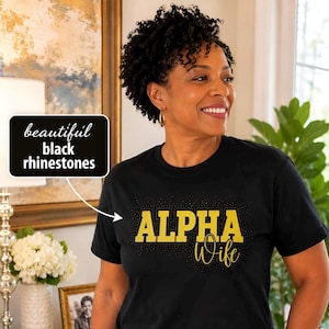 May include: A black short-sleeved t-shirt featuring the text ALPHA Wife in yellow lettering. The design is accented with black rhinestones. A black rectangular callout box with white text reads beautiful black rhinestones.