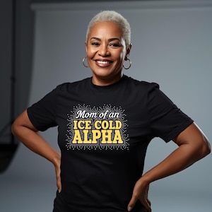 May include: Black t-shirt with the text "Mom of an ICE COLD ALPHA" in white and yellow. The text is embellished with small white dots. The woman in the image is smiling and wearing gold hoop earrings.