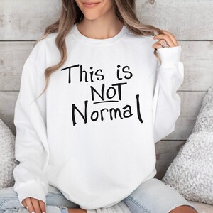 May include: White sweatshirt with the phrase "This is NOT Normal" in black lettering. The sweatshirt is a crew neck style and is being worn.