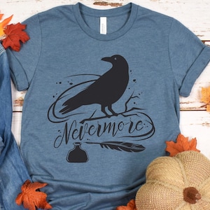May include: A blue t-shirt with a black silhouette of a raven perched on a branch. The text "Nevermore" is written in a stylized font below the raven. A black inkwell and quill are also depicted.