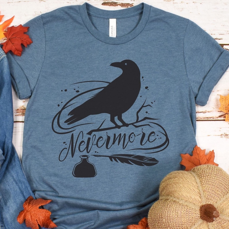 May include: A blue t-shirt with a black silhouette of a raven perched on a branch. The text "Nevermore" is written in a stylized font below the raven. A black inkwell and quill are also depicted.