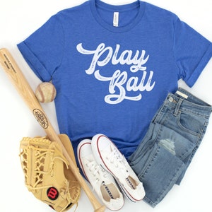 Peut inclure: Un t-shirt bleu avec l'inscription "Play Ball" en lettres blanches vieillies. Le t-shirt est associ&eacute; &agrave; une batte de baseball, un gant de baseball, une balle de baseball et une paire de baskets blanches.