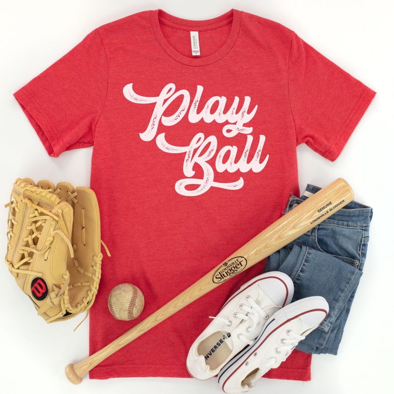 Peut inclure: T-shirt rouge avec un texte blanc qui dit "Play Ball" dans une police de caract&egrave;res vintage. Le t-shirt est pos&eacute; sur une surface blanche avec une batte de baseball, un gant de baseball, une balle de baseball et des baskets blanches.