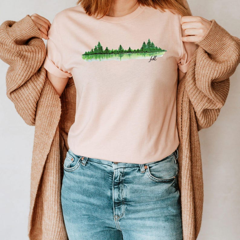 May include: A pink t-shirt with a watercolor design of a forest reflected in a lake. The text "Felt" is printed below the design.