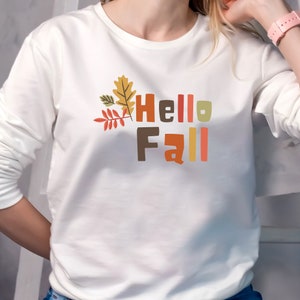 May include: White long-sleeved shirt with a graphic print of colorful leaves and the text "Hello Fall".
