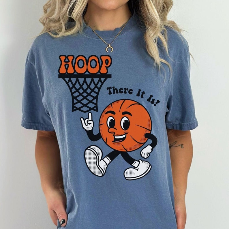 May include: A blue t-shirt with a cartoon basketball going through a hoop. The basketball has a face and is saying "There It Is!"