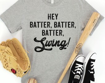 May include: A gray t-shirt with the text "Hey Batter, Batter, Batter, Swing!" printed on it. A baseball bat, baseball glove, and baseball are also in the image.