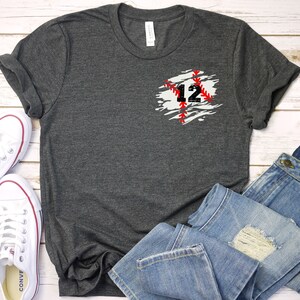 K&ouml;nnte beinhalten: Dunkelgraues T-Shirt mit Baseball-Design. Das Design zeigt die Zahl 12 in Schwarz mit roten N&auml;hten auf einem wei&szlig;-grauen Hintergrund. Das Shirt wird mit blauen Jeans und wei&szlig;en Turnschuhen gezeigt.