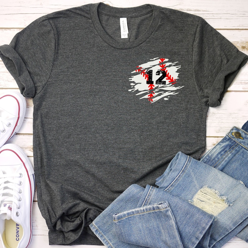 K&ouml;nnte beinhalten: Dunkelgraues T-Shirt mit Baseball-Design. Das Design zeigt die Zahl 12 in Schwarz mit roten N&auml;hten auf einem wei&szlig;-grauen Hintergrund. Das Shirt wird mit blauen Jeans und wei&szlig;en Turnschuhen gezeigt.