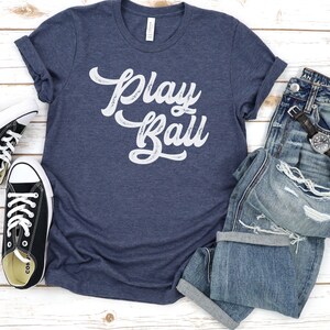 Peut inclure: Un t-shirt bleu marine avec le texte "Play Ball" en blanc, lettres vieillies. Le t-shirt est pos&eacute; &agrave; plat sur une surface en bois blanc avec une paire de baskets noires et un jean bleu.