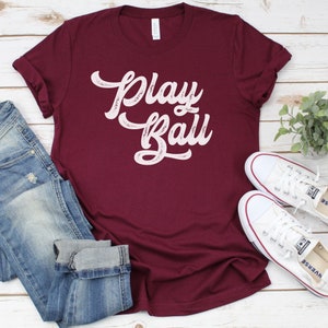 Peut inclure: Un t-shirt bordeaux avec les mots "Play Ball" en &eacute;criture blanche sur le devant. Le t-shirt est pos&eacute; sur une surface en bois blanc avec une paire de chaussures en toile blanche et un jean bleu.