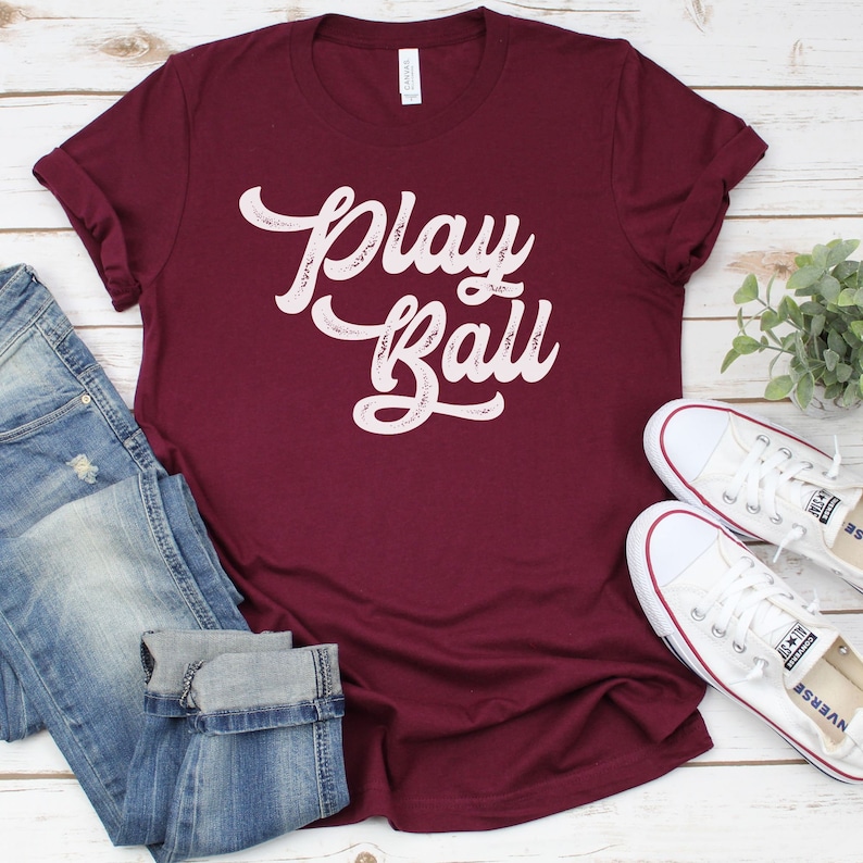 Peut inclure: Un t-shirt bordeaux avec les mots "Play Ball" en &eacute;criture blanche sur le devant. Le t-shirt est pos&eacute; sur une surface en bois blanc avec une paire de chaussures en toile blanche et un jean bleu.